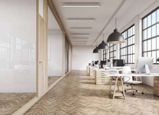 Vista frontal del interior de una oficina con una fila de mesas de madera bajo grandes ventanales. Lámparas masivas en el plafón. Computadoras, 3d rendering vía Shutterstock