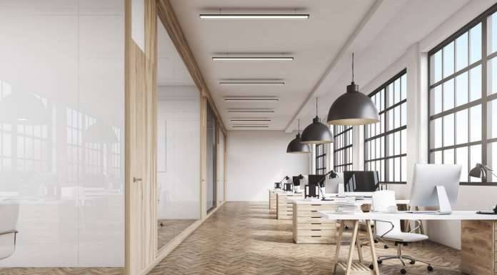 Vista frontal del interior de una oficina con una fila de mesas de madera bajo grandes ventanales. Lámparas masivas en el plafón. Computadoras, 3d rendering vía Shutterstock