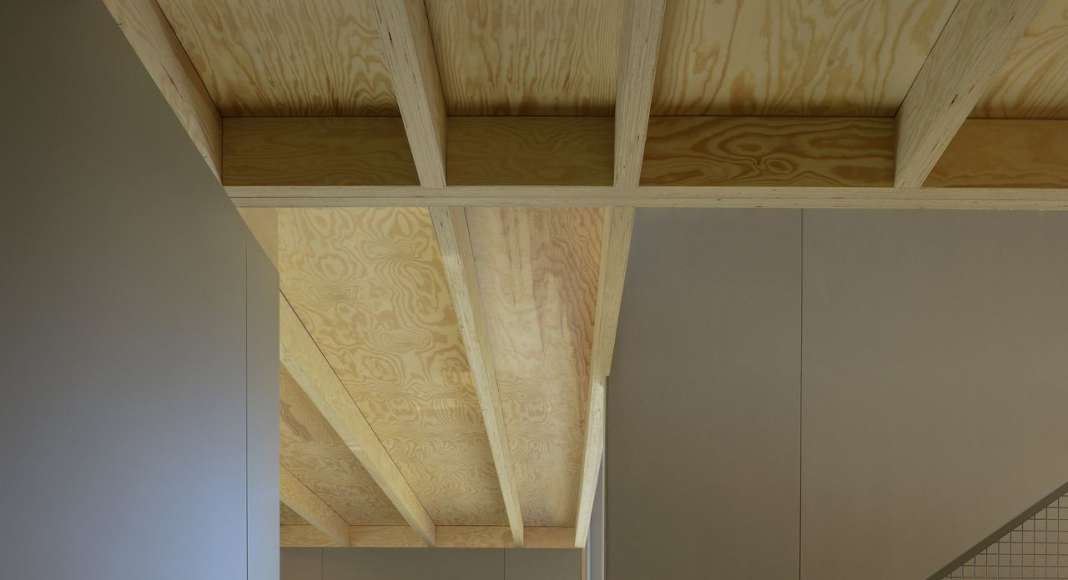 Interior view from guest room to staircase and kitchen. The staircase is made of pine with a sliding rack attached to the catch. The roof with visible solid wood construction and walls of grey coloured wood fibreboards. The floor is glossy and dust-bound concrete floor. : Photo © Bornstein Lyckefors Arkitekter