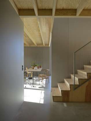 Interior view from guest room to staircase and kitchen. The staircase is made of pine with a sliding rack attached to the catch. The roof with visible solid wood construction and walls of grey coloured wood fibreboards. The floor is glossy and dust-bound concrete floor. : Photo © Bornstein Lyckefors Arkitekter